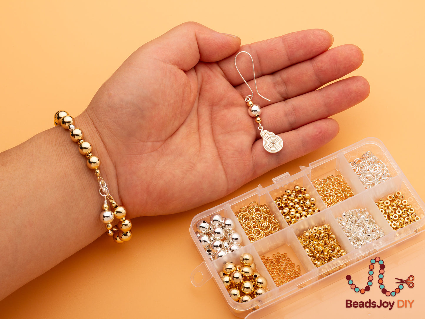 Hand holding a completed gold bead bracelet and handmade wire-wrapped earring next to a 10-grid storage box filled with gold and silver beads and findings