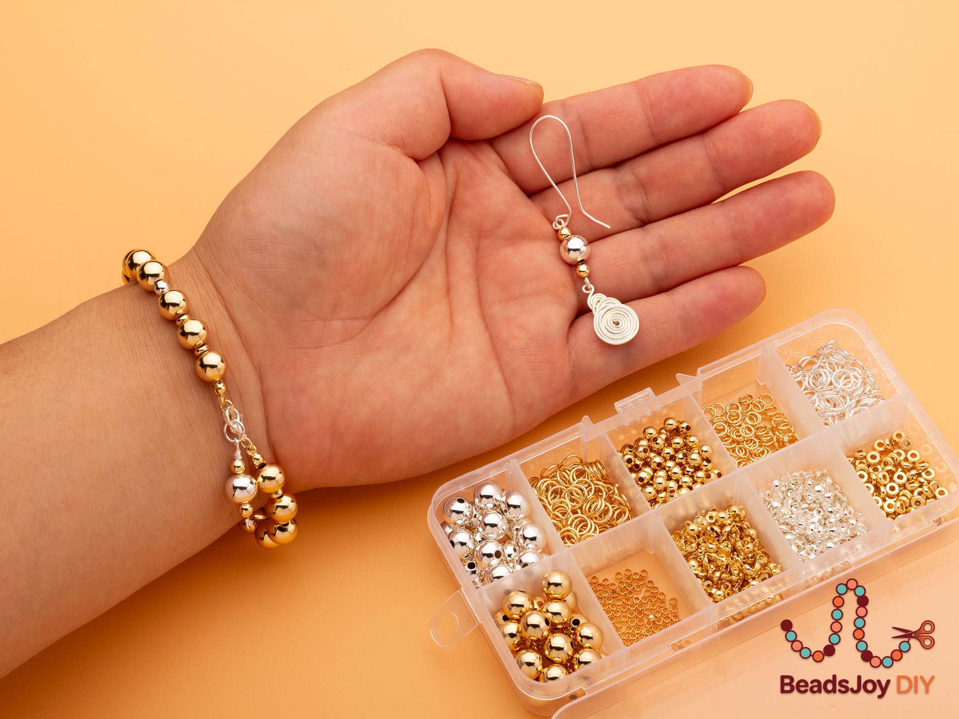 Hand holding a completed gold bead bracelet and handmade wire-wrapped earring next to a 10-grid storage box filled with gold and silver beads and findings