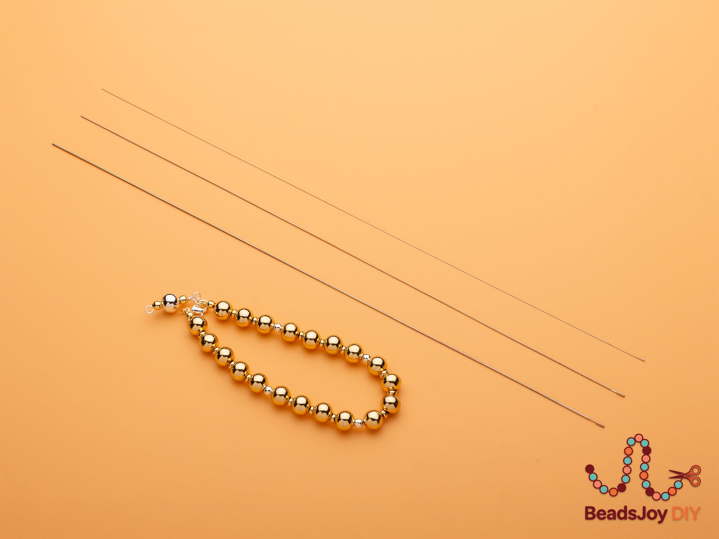 Completed gold bead bracelet with lobster clasp and three beading wires laid flat on orange background – BeadsJoy DIY